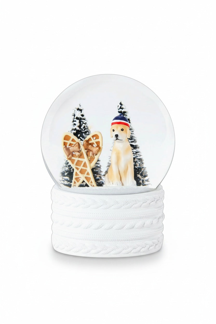 A ZODAX SNOW SHOEING MOUNTAIN DOG SNOW GLOBE featuring a resin glass globe with a dog in a hat, snowshoes, and two pine trees, set on a white cable-knit textured base.