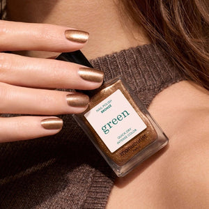 A hand with shimmery bronze vegan, cruelty-free nail polish holds a matching bottle labeled “MANUCURIST - GREEN™ Nail Polish in Tanned” against a brown sweater.