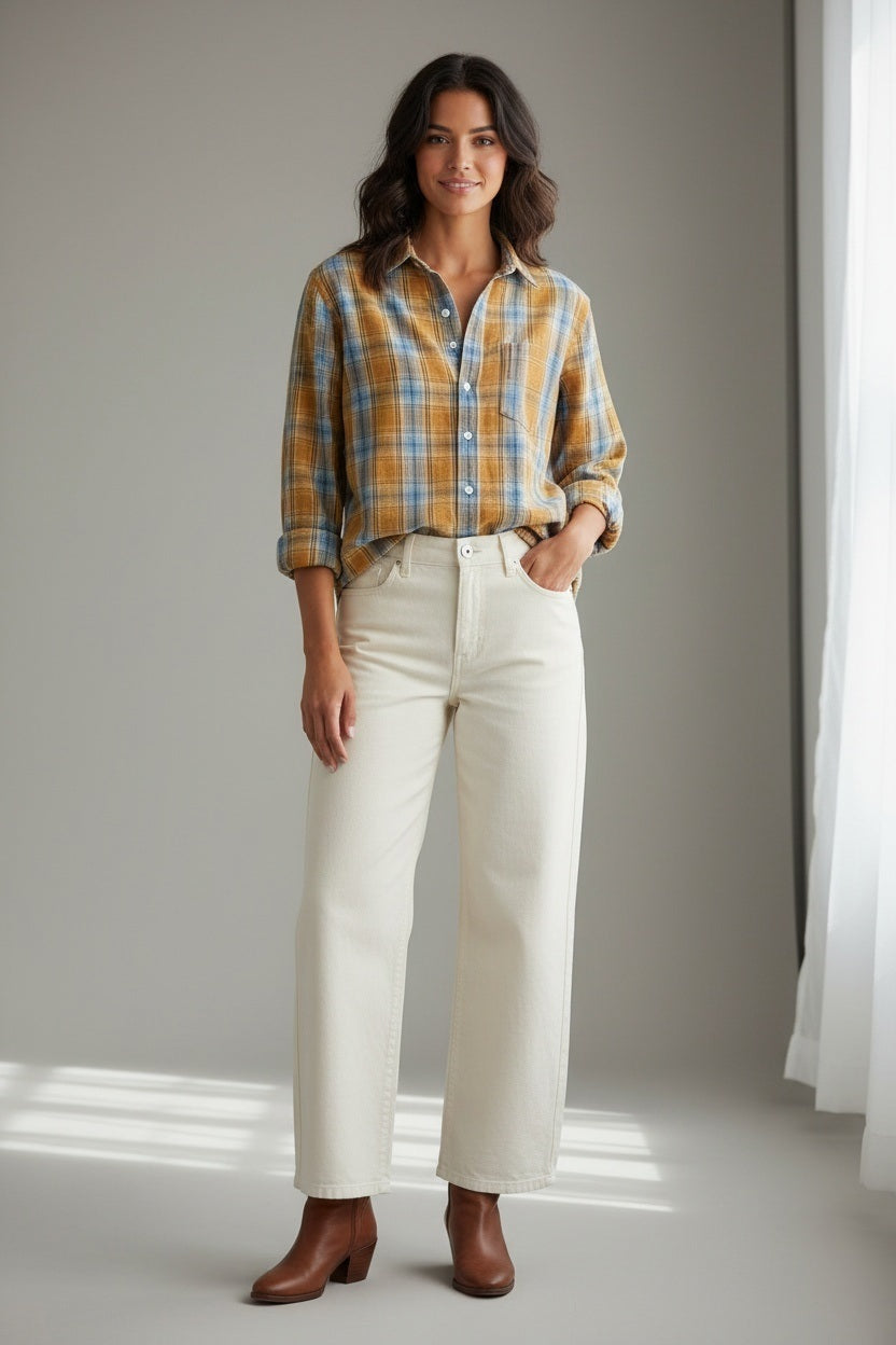 A woman stands indoors against a neutral background with soft light streaming in, smiling gently with one hand in her pocket. She wears the FRANK & EILEEN Eileen Relaxed Button Up Shirt in Italian Flannel Gold Blue Large Plaid—featuring a bust-flattering button placement—paired with cream high-waisted pants and brown ankle boots.