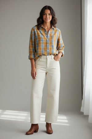 A woman stands indoors against a neutral background with soft light streaming in, smiling gently with one hand in her pocket. She wears the FRANK & EILEEN Eileen Relaxed Button Up Shirt in Italian Flannel Gold Blue Large Plaid—featuring a bust-flattering button placement—paired with cream high-waisted pants and brown ankle boots.