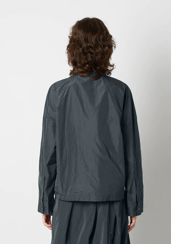 A person with shoulder-length wavy hair is wearing the KATHARINA HOVMAN Kimono Blouse from the brand KATHARINA HOVMAN. The blouse is long-sleeved, button-up, and dark gray. They are standing against a plain light gray background, gazing slightly upward with a calm expression while lightly clasping their hands in front.