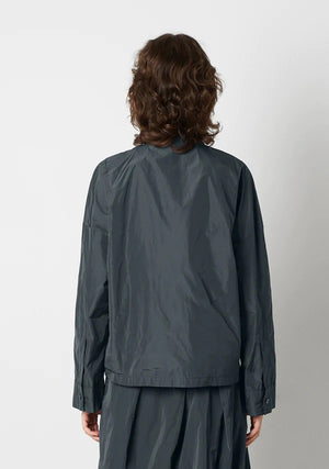 A person with shoulder-length wavy hair is wearing the KATHARINA HOVMAN Kimono Blouse from the brand KATHARINA HOVMAN. The blouse is long-sleeved, button-up, and dark gray. They are standing against a plain light gray background, gazing slightly upward with a calm expression while lightly clasping their hands in front.