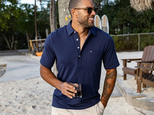 A smiling man in sunglasses stands outside by a wooden building, holding a drink and wearing the CRIQUET Top-Shelf Players Shirt in Navy. Sunlight and greenery are visible in the background.