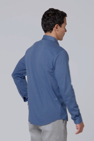 A man with short dark hair wears the MACK WELDON Silver Pique Long Sleeve Button Up Shirt in True Navy, standing against a plain light background with a neutral expression and his hands together in front of him.