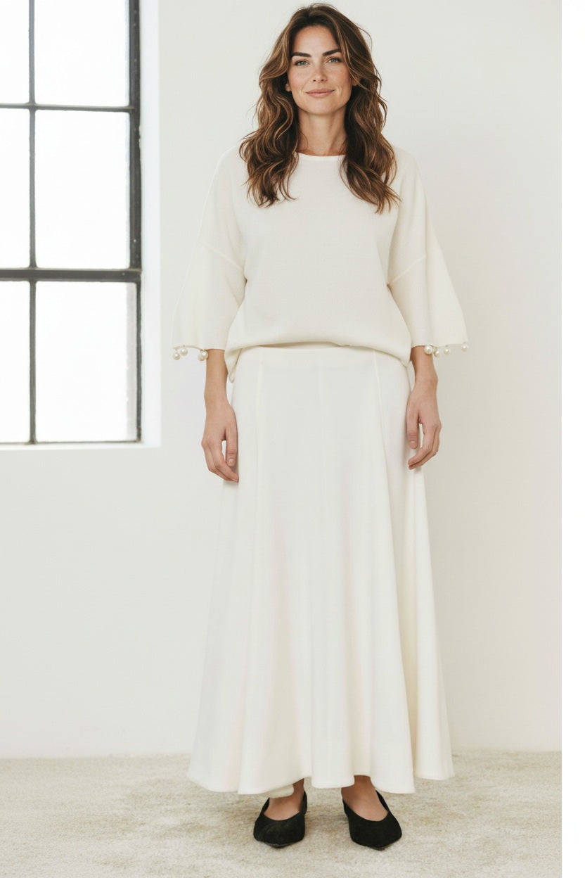 A person wears the MEIMEIJ - SKIRT, a long, flowing minimalist piece by MEIMEIJ, styled with a white top with pearl-detailed sleeves and black flats, standing on a light carpet.