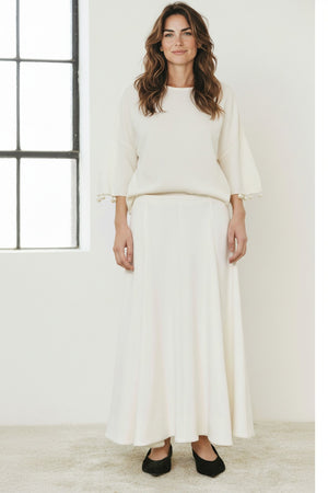 A person wears the MEIMEIJ - SKIRT, a long, flowing minimalist piece by MEIMEIJ, styled with a white top with pearl-detailed sleeves and black flats, standing on a light carpet.