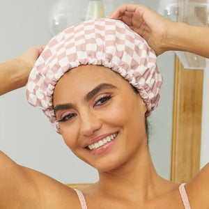 The KITSCH - SATIN LINED FLEXI SHOWER CAP, featuring a checkered pink and white design, is waterproof with satin lining and comes with adjustable closure for various hair types, as shown on its packaging.