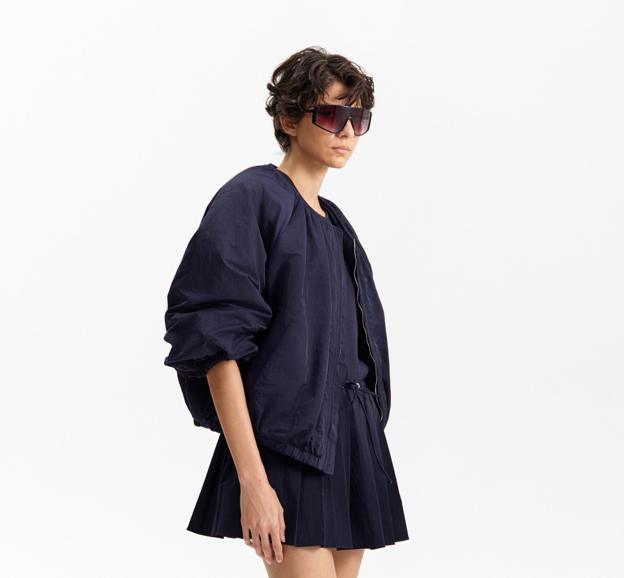A person models the ELEH NYLON BOMBER JACKET, styled oversized with a matching mini skirt, black ankle boots, and large sunglasses against a plain white background. Brand: ELEH.