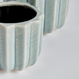 The NAPA HOME AND GARDEN - VELA POT, SMALL is a light blue ceramic decorative flower pot featuring vertical ridges and a cactus-inspired design, shown in a side view with part of another planter visible in the background.