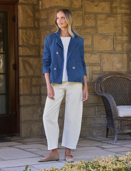 A woman stands smiling in front of a stone wall, wearing the FRANK & EILEEN Mini Belfast Cropped English Peacoat in 1984 Wash Denim over a white top and white pants.