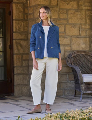 A woman stands smiling in front of a stone wall, wearing the FRANK & EILEEN Mini Belfast Cropped English Peacoat in 1984 Wash Denim over a white top and white pants.