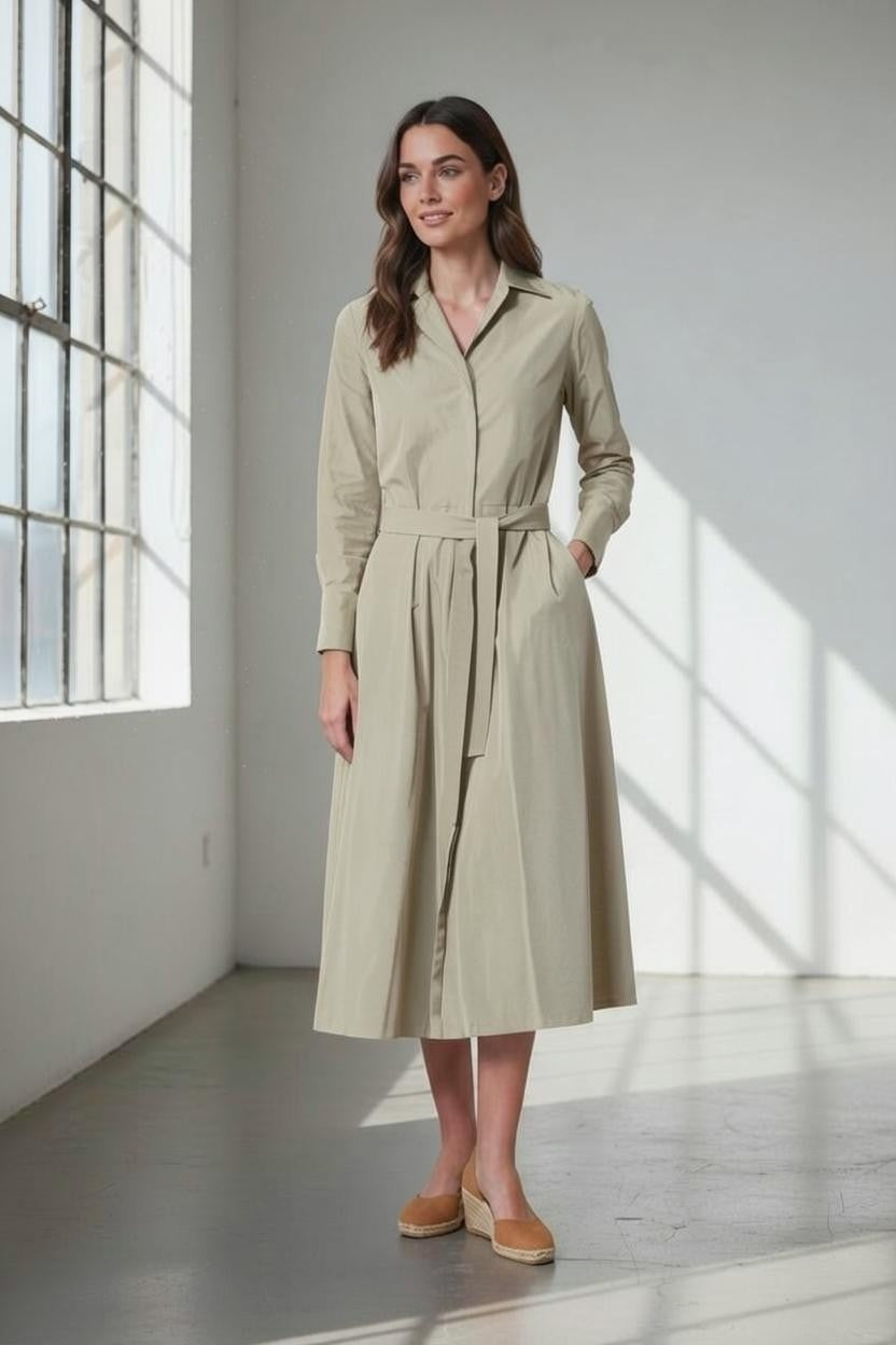 A woman stands in a sunlit room, her hands in her pockets and a slight smile on her face, wearing the VICARIO CINQUE ANNA Taffeta Shirtdress in Light Taupe.