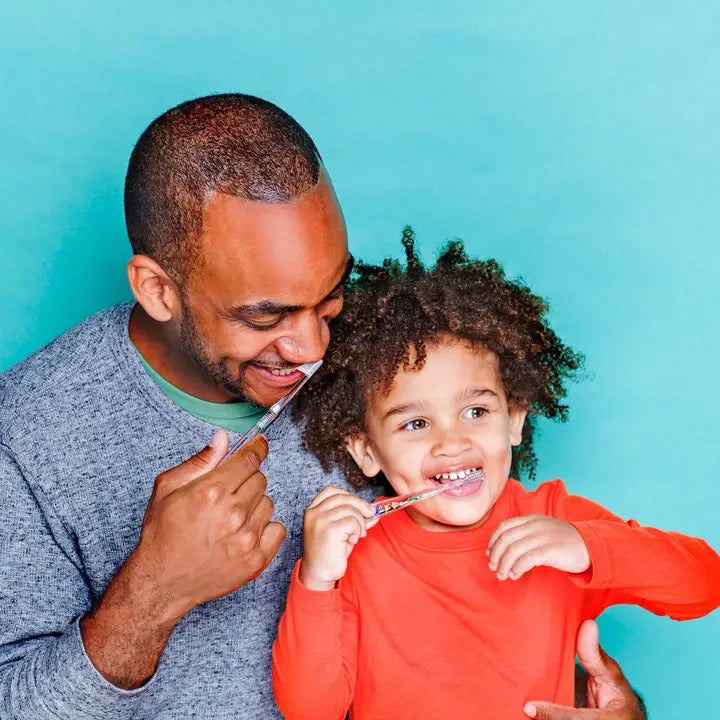Two HAMICO - KIDS DINO TOOTHBRUSHES by HAMICO/INNOVATIVE TOOTHBRUSH, featuring unique dinosaur designs, sit on a white countertop next to a beige towel. A hint of a green plant is visible in the upper right corner, adding a touch of natural decor to the scene.