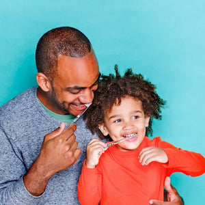Two HAMICO - KIDS DINO TOOTHBRUSHES by HAMICO/INNOVATIVE TOOTHBRUSH, featuring unique dinosaur designs, sit on a white countertop next to a beige towel. A hint of a green plant is visible in the upper right corner, adding a touch of natural decor to the scene.