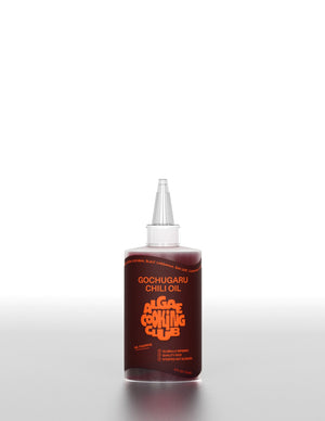 A clear plastic bottle labeled "ALGAE COOKING CLUB - GOCHUGARU CHILI OIL" in bold orange text sits on a white surface against a plain background, holding vibrant Korean chili oil from ALGAE COOKING CLUB.