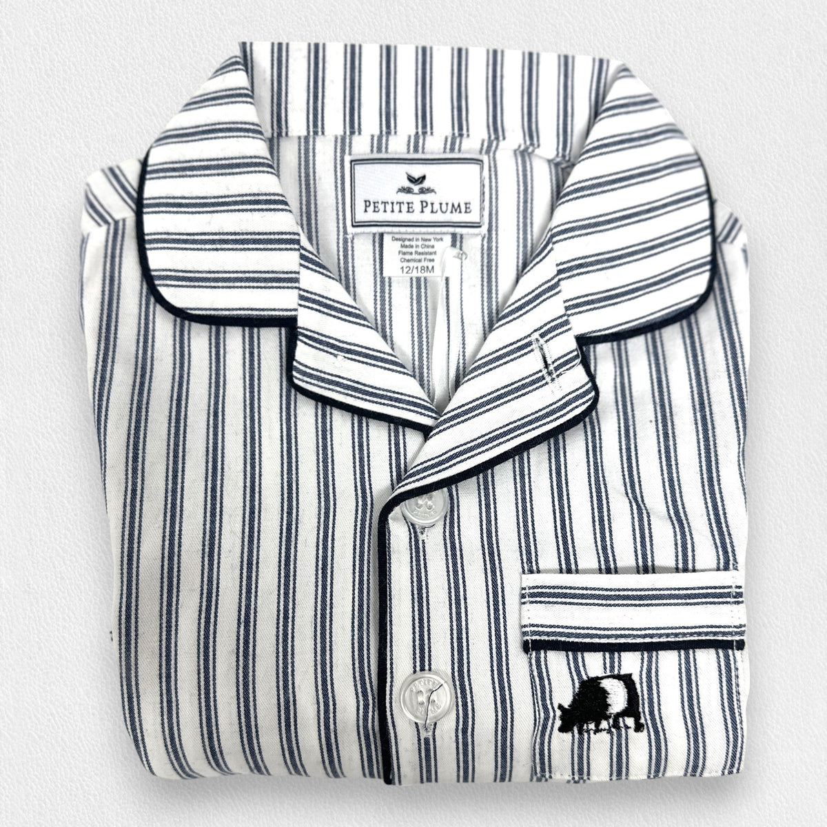 The PETITE PLUME FEARRINGTON LIFESTYLE COLLECTION - CHILDREN&#39;S TWILL NAVY FRENCH TICKING PAJAMA SET is a delightful option for kids, featuring long sleeves and pants with chic vertical white and gray stripes. This set is crafted from yarn-dyed cotton, boasting a top with a collar, full button-down front, and a single chest pocket adorned with matching fabric piping along the cuffs and pocket opening.
