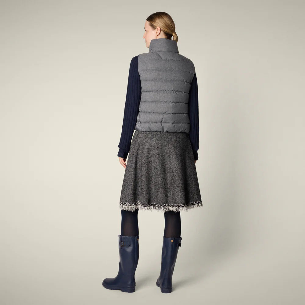 A woman with her hair tied back smiles against a plain background, wearing the SAVE THE DUCK - NYLA PUFFER VEST by SAVE THE DUCK over a navy turtleneck, gray fringed A-line skirt, navy tights, and blue rain boots.