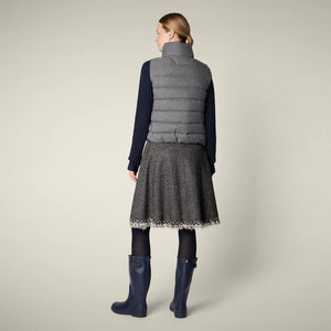 A woman with her hair tied back smiles against a plain background, wearing the SAVE THE DUCK - NYLA PUFFER VEST by SAVE THE DUCK over a navy turtleneck, gray fringed A-line skirt, navy tights, and blue rain boots.