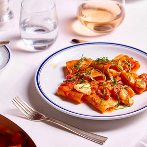 A plate of Italian pasta made from organic durum wheat semolina, such as CIPRIANI - ORGANIC PACCHERI DURUM WHEAT PASTA by ATALANTA CORPORATION, with tomato sauce and basil sits on a white tablecloth next to a fork, a glass of water, and a glass of rosé wine.