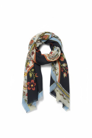 The STORIATIPIC ARIANE wool scarf with embroidery is adorned with a floral and vine pattern in red, green, blue, cream, and black tones. It features light blue edges and a soft, slightly fringed border. The embroidered scarf is loosely knotted against a white background.