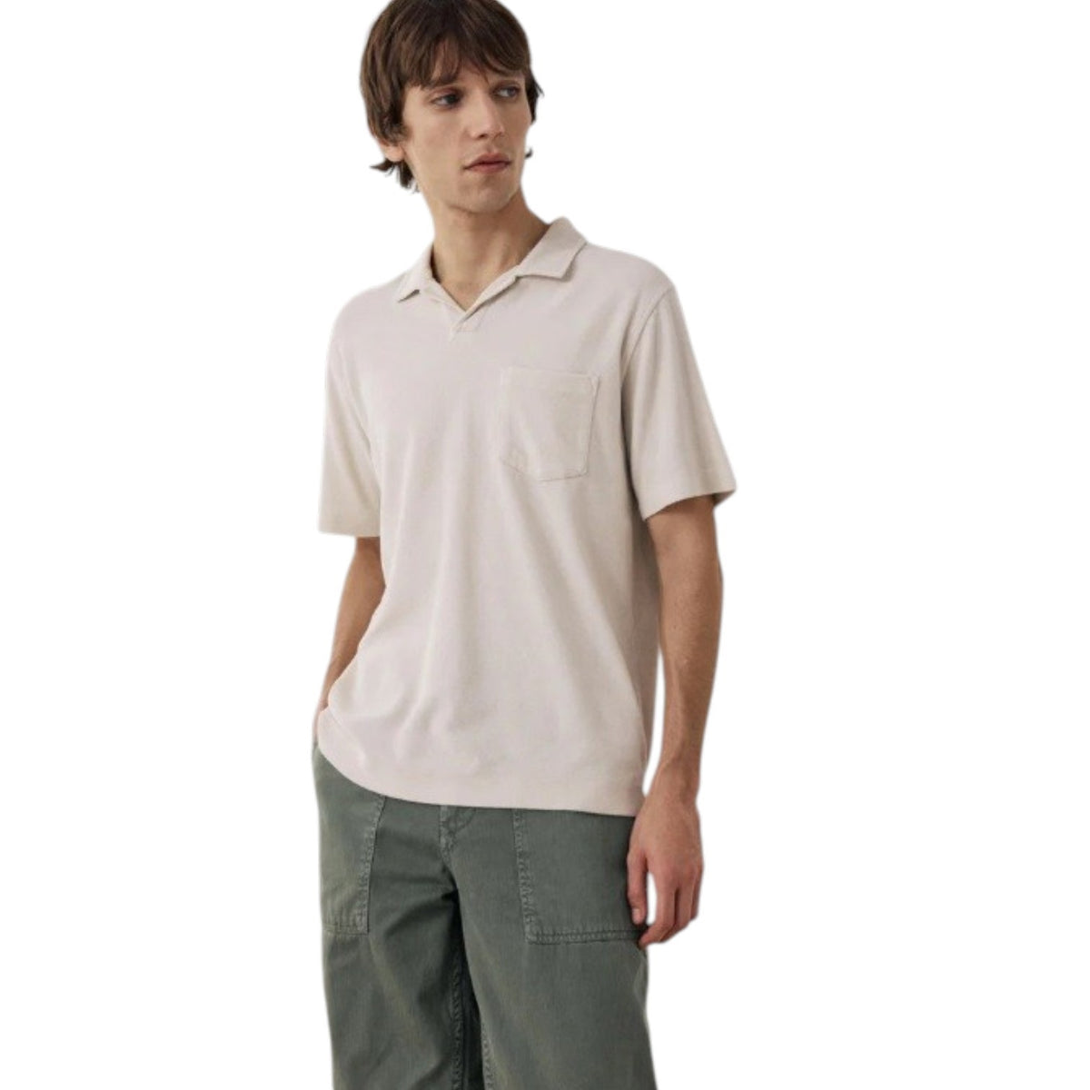Wearing the HARTFORD Terry Polo Shirt in Stone with sage green cargo pants, a person stands with one hand in their pocket, glancing to the side against a plain background.