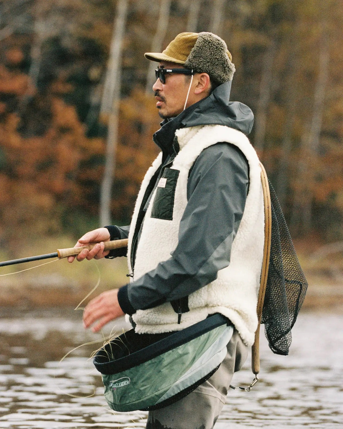 A man in the AMUNDSEN SPORTS - HARVESTER REVERSO WAXED COTTON VEST and sunglasses stands in shallow water with a fishing rod and net, surrounded by autumn trees.