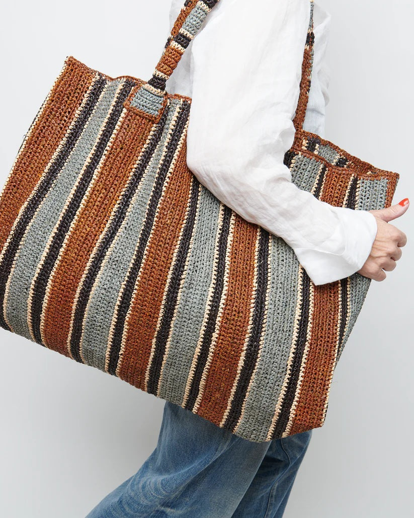 A woman stands on a sunlit path in a sleeveless dress and long scarf, holding the GABRIELE FRANTZEN - RAFFIA WEEKENDER BAG TOBACCO. She looks over her shoulder, surrounded by lush greenery.