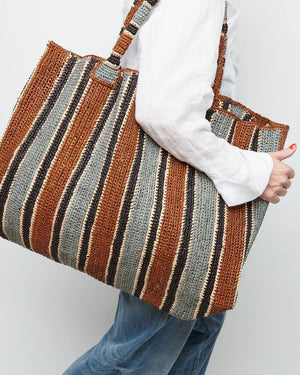 A woman stands on a sunlit path in a sleeveless dress and long scarf, holding the GABRIELE FRANTZEN - RAFFIA WEEKENDER BAG TOBACCO. She looks over her shoulder, surrounded by lush greenery.