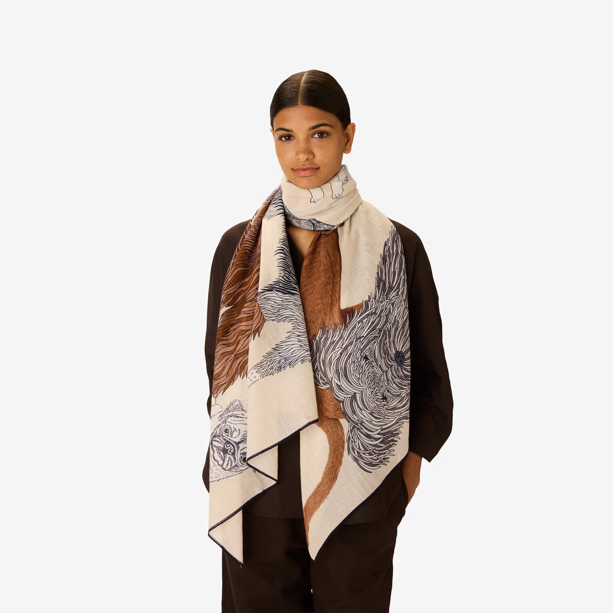 A woman in a dark outfit stands with her hands in her pockets, draped in the INOUI EDITIONS - TOUTOU Scarf in White by INOUI, featuring intricate dark brown and navy animal illustrations. She is set against a plain white background.