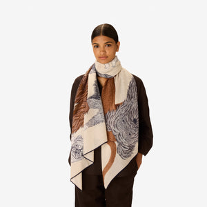 A woman in a dark outfit stands with her hands in her pockets, draped in the INOUI EDITIONS - TOUTOU Scarf in White by INOUI, featuring intricate dark brown and navy animal illustrations. She is set against a plain white background.