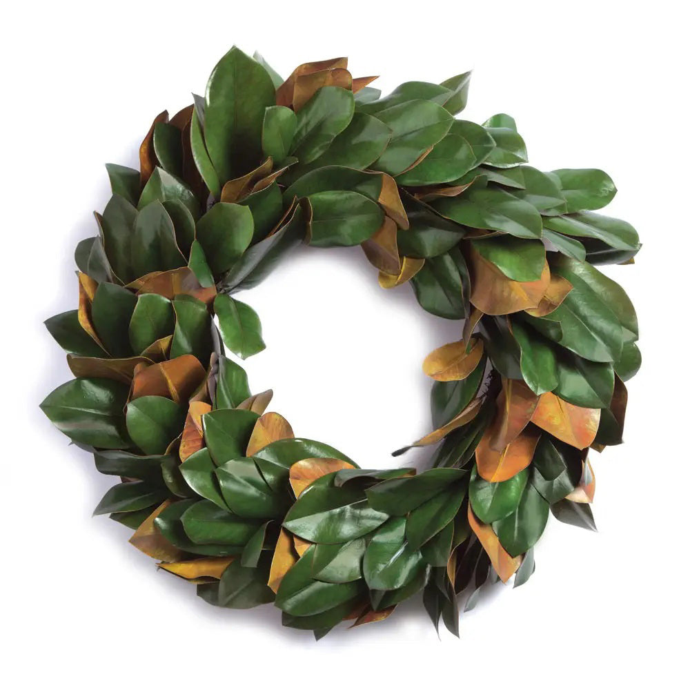 A lush green FAUX MAGNOLIA WREATH 24" from NAPA HOME AND GARDEN adorns a white, ornate fireplace mantel set against a brick surround. Below, a gentle fire burns inside the fireplace, adding warmth to the room's elegant and cozy atmosphere—perfect for interior use as a captivating centerpiece.