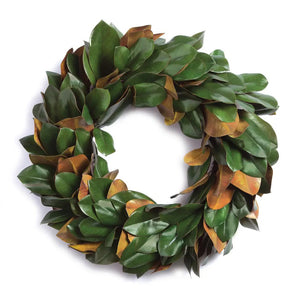 A lush green FAUX MAGNOLIA WREATH 24" from NAPA HOME AND GARDEN adorns a white, ornate fireplace mantel set against a brick surround. Below, a gentle fire burns inside the fireplace, adding warmth to the room's elegant and cozy atmosphere—perfect for interior use as a captivating centerpiece.