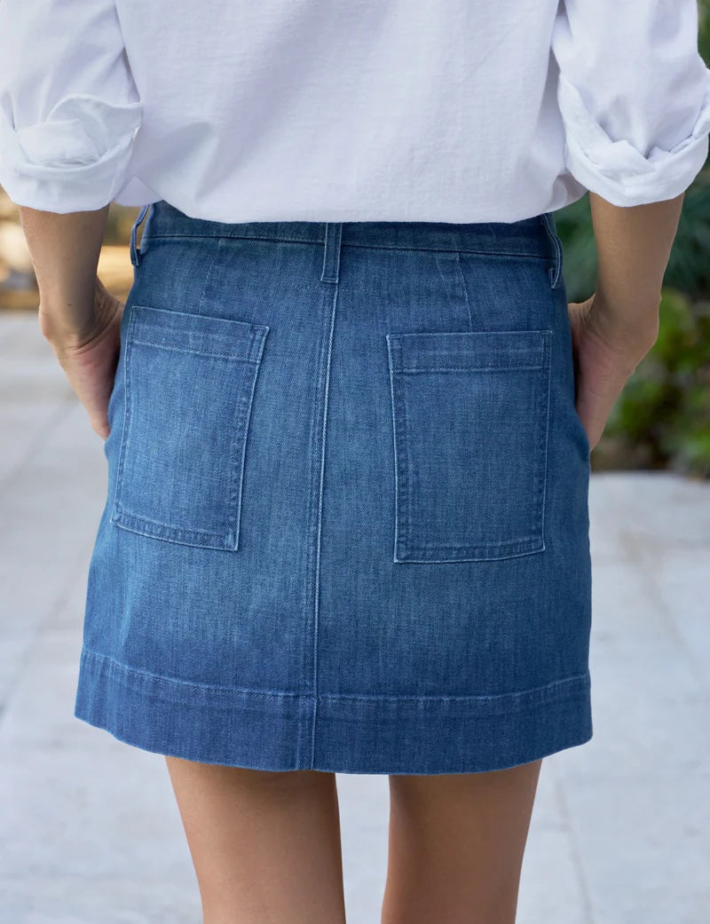 Close-up of a person wearing the FRANK &amp; EILEEN Jameson Utility Skirt in Italian Dream Light Denim 1984 Wash, with utility pockets, a white shirt, and a gold ring. Their right hand rests in the front pocket; background is blurred.
