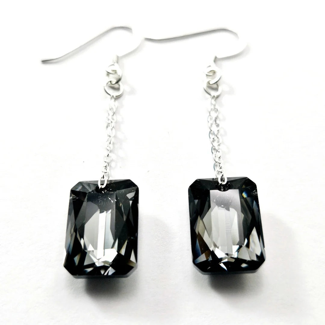 Introducing the VERDIER - SWAROVSKI NIGHT CRYSTAL EARRINGS by VERDIER: a pair of sterling silver drop earrings featuring rectangular, emerald-cut Swarovski crystals suspended from delicate chains, beautifully showcased against a white background.