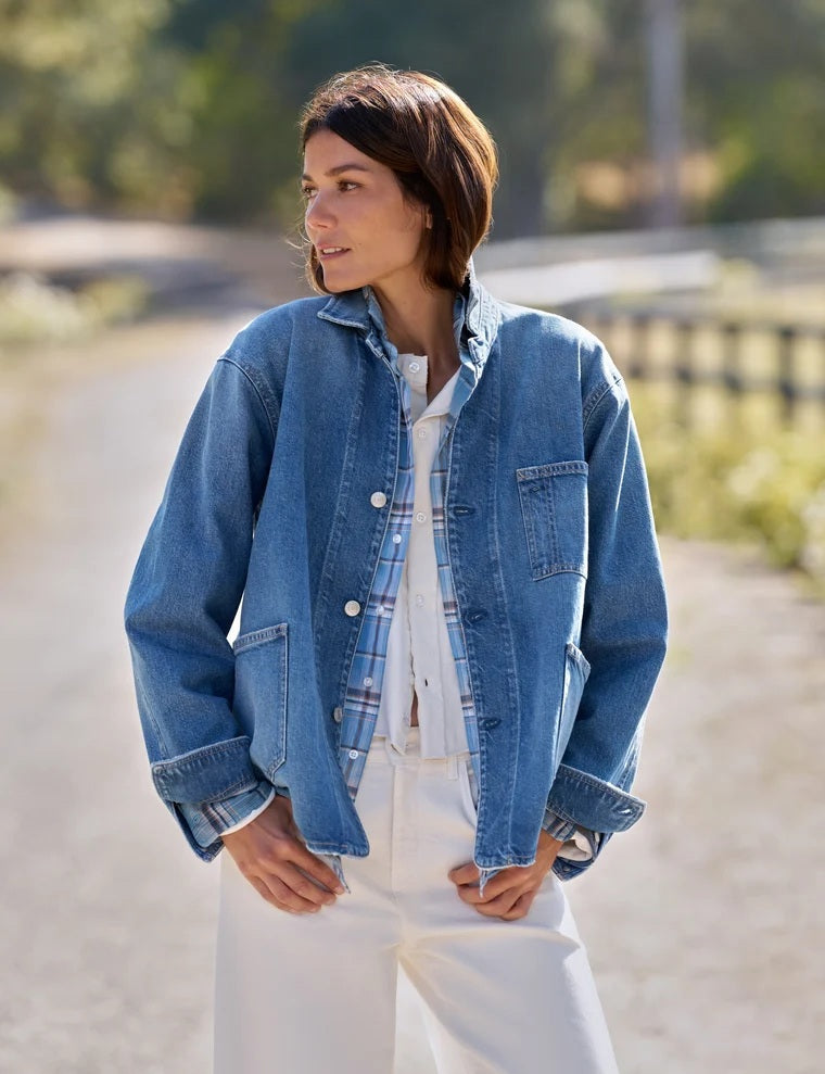A woman stands indoors wearing the FRANK & EILEEN Bantry Barn Jacket in California Vintage Denim 1978 Wash over a white top, blue jeans, and brown ankle boots. She has short, dark hair and is smiling as natural light streams through a large window beside her.