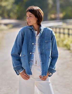 A woman stands indoors wearing the FRANK & EILEEN Bantry Barn Jacket in California Vintage Denim 1978 Wash over a white top, blue jeans, and brown ankle boots. She has short, dark hair and is smiling as natural light streams through a large window beside her.