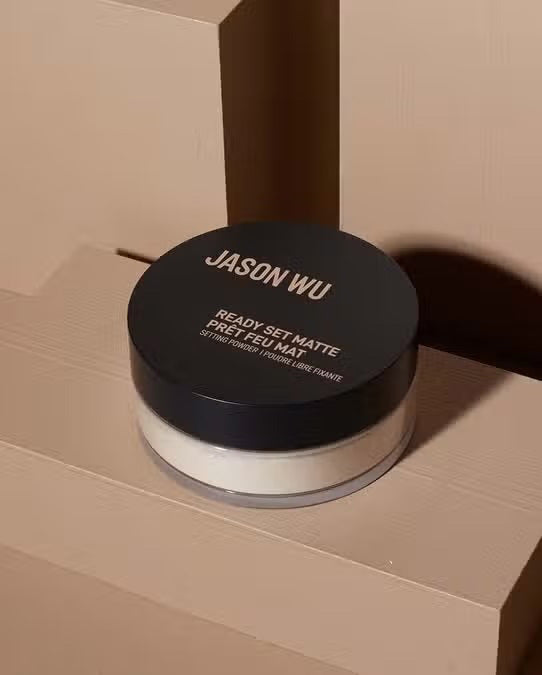 A small round container of JASON WU BEAUTY - READY SET MATTE 2 translucent setting powder is placed on a light brown, geometric background. The container features a black lid with white text displaying the product name, promising a flawless matte finish.