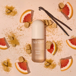 A bottle of KITSCH - HAIR PERFUME MIST IN WARM SUGAR stands between a slice of cake and blue cotton candy, with labeled fragrance notes: pink grapefruit, whipped cream, spun sugar, bergamot blossom, musk, vanilla, and praline. This odor-eliminating formula from KITSCH ensures your hair smells irresistibly sweet and fresh all day.