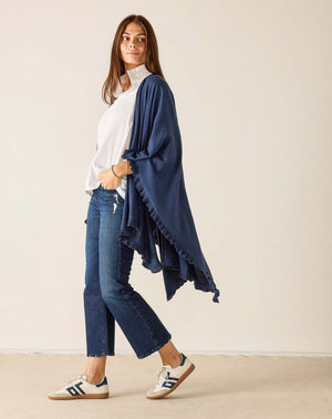 A woman with long brown hair sits sideways on a wooden chair, wearing the MERSEA - CHARLESTON RUFFLE WRAP IN CHAMBRAY over a white shirt, jeans, and sneakers, looking at the camera against a plain background.