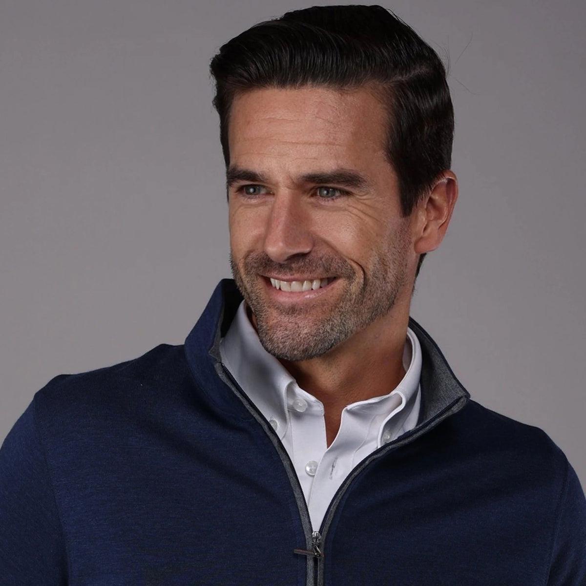 A man with short brown hair and a beard smiles while wearing the COLLARS AND CO - Heather Cotton Cloud 1/4 Zip Sweater in Navy with Grey Trim over a white collared shirt and cream pants, standing against a plain gray background.