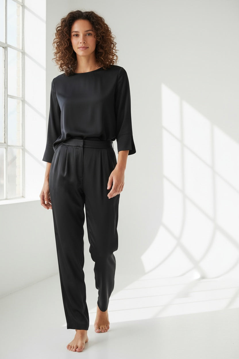 A person is wearing HINSON WU’s HOSTESS LIQUID SATIN PANT IN BLACK—high-waisted, wide-leg pants crafted from lustrous liquid satin fabric—paired with a black three-quarter sleeve top and open-toe heels with a thin strap. Their left hand is in the pocket of the pants, and their head is not visible.