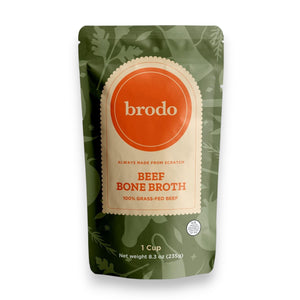 A pouch of BRODO BROTH- GRASS FEED BONE BROTH from BRODO BROTH COMPANY rests on a wooden surface, accompanied by a cup of collagen-rich broth, celery sticks, salt crystals, black peppercorns, and a bay leaf—ideal for sipping or crafting a rich pan sauce.