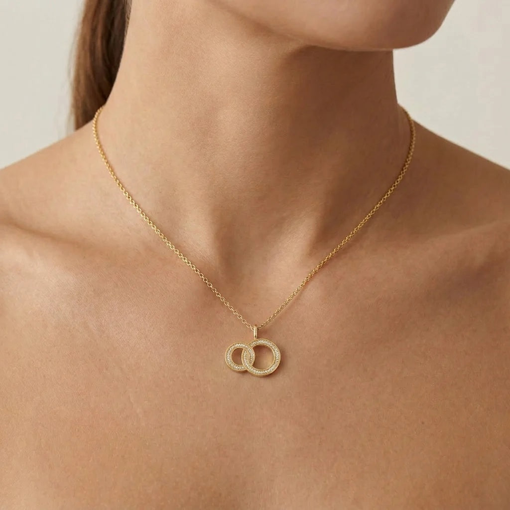 The ELLA STEIN - FOREVER LINKED PENDANT NECKLACE by ELLA STEIN features two interlocking circle pendants, each adorned with sparkling gemstones, suspended from a delicate gold-plated stainless steel chain and displayed against a white background.