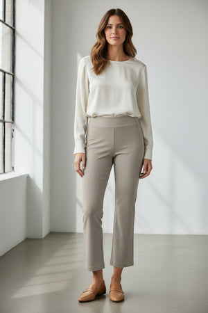 A woman stands indoors by a large window, wearing a long-sleeve cream blouse and the KRAZY LARRY, INC PULL ON WIDE ANKLE PANT in black, which features a tummy tuck design. She completes her look with black loafers and smiles slightly, her wavy brown hair catching the sunlight that casts shadows on the white floor and wall.
