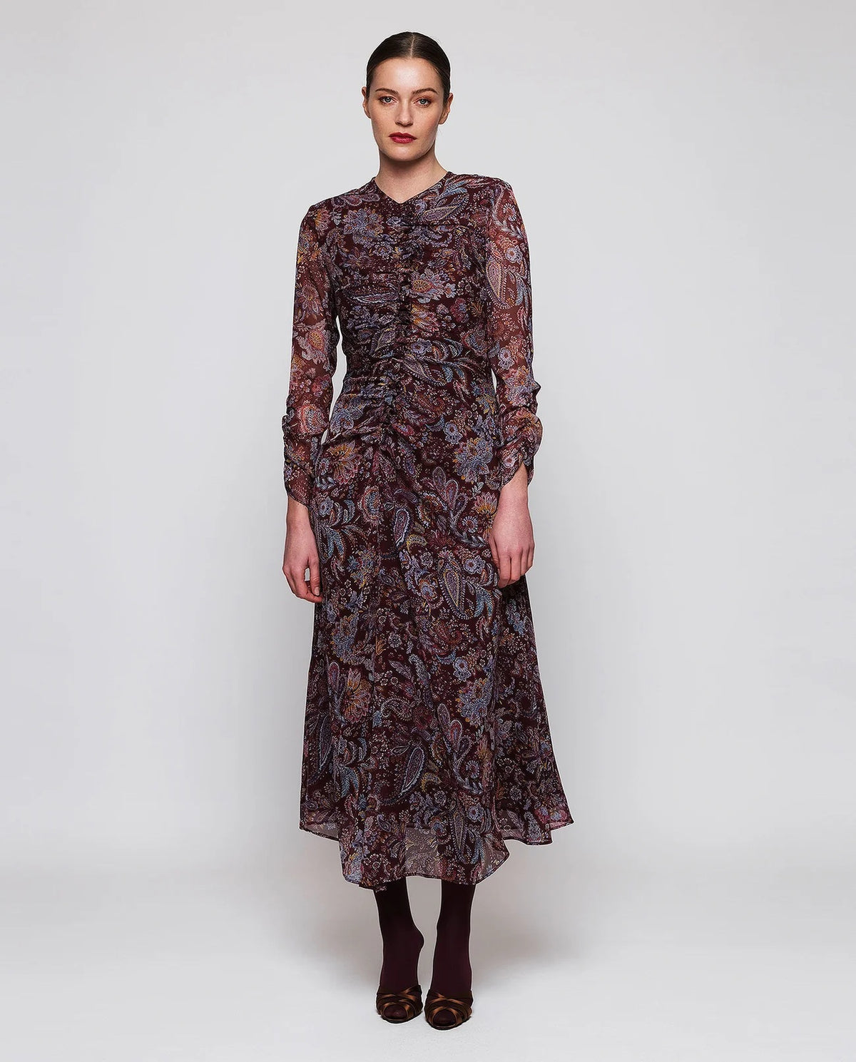 A woman stands facing away, wearing the MIRTO 1956 Burgundy Draped Paisley Print Dress—a long sleeve midi dress featuring an elegant paisley pattern—paired with burgundy tights and black heels. Her hair is styled in a neat bun as she poses against a plain white background.