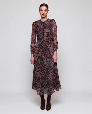 A woman stands facing away, wearing the MIRTO 1956 Burgundy Draped Paisley Print Dress—a long sleeve midi dress featuring an elegant paisley pattern—paired with burgundy tights and black heels. Her hair is styled in a neat bun as she poses against a plain white background.