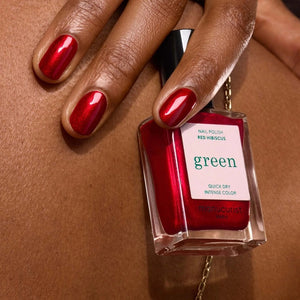 A hand with shiny, iridescent red nails holds a matching bottle labeled “MANUCURIST - GREEN™ NAIL POLISH IN RED HIBISCUS” by MANUCURIST INC against brown skin. This luxurious vegan, cruelty-free nail polish delivers vibrant color and a flawless finish.