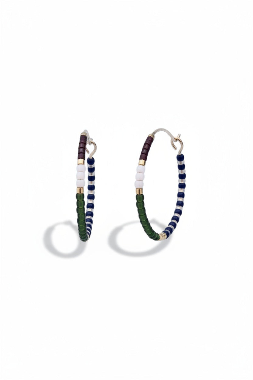 The ALICE RISE - MIDI COLORLOOP EARRINGS IN OLIVE by ALICE RISE feature 14k gold filled hoops adorned with white, navy blue, green, burgundy, and gold Japanese glass beads, displayed on a textured light blue surface.