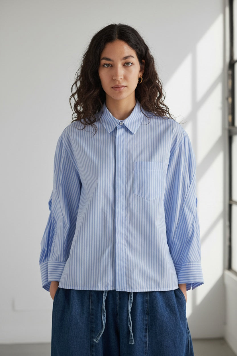 A woman with dark hair tied back models the MEIMEIJ STRIPE COTTON SHIRT—an oversized light blue and white striped cotton top with wide sleeves—paired with blue pants, standing against a plain light gray background.