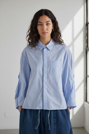 A woman with dark hair tied back models the MEIMEIJ STRIPE COTTON SHIRT—an oversized light blue and white striped cotton top with wide sleeves—paired with blue pants, standing against a plain light gray background.